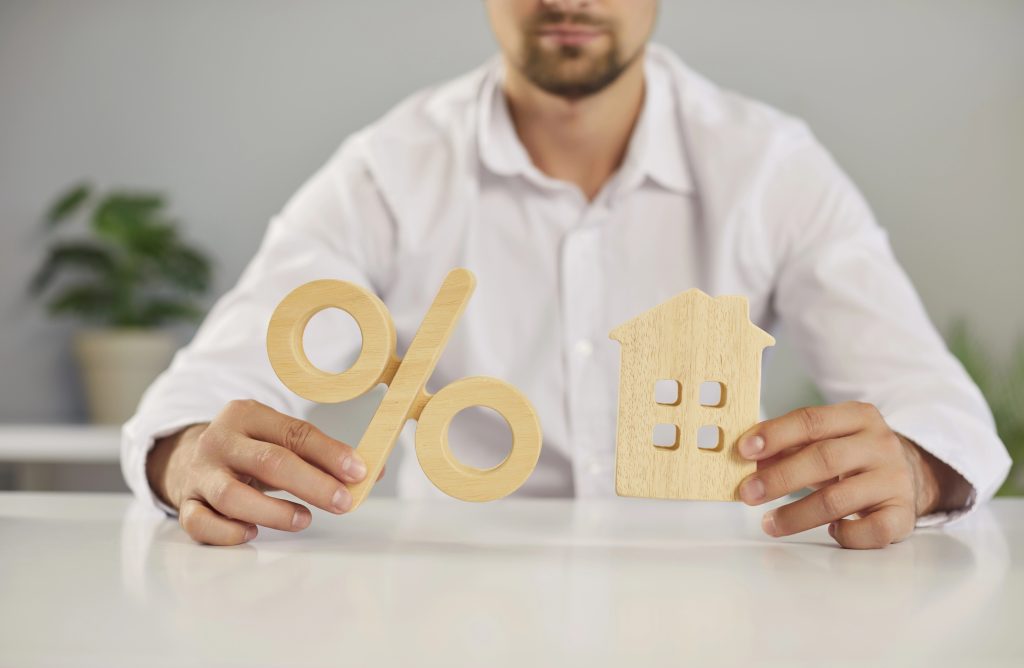 Cropped shot of a male real estate agent sitting at a table and holding a wooden percent sign and a small toy house. Concept of a mortgage interest rate