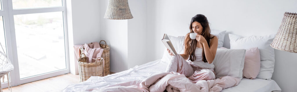 Young woman with cup of coffee reading book on bed, banner